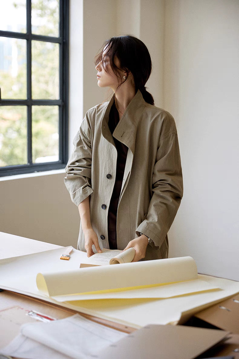 Woman wearing the gray Relaxed Cotton-Linen Trench standing by a desk near the window, highlighting the relaxed shape, button front, and breathable fabric