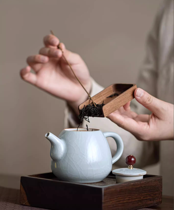 Ru Kiln Celadon Mini Teapot in light blue with lid removed as loose tea leaves are added using a wooden scoop on a tea tray
