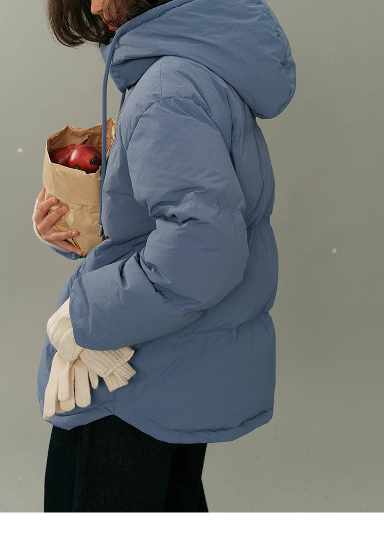 Side-angle view of the dusty blue puffer jacket as the model holds a pear and looks away.