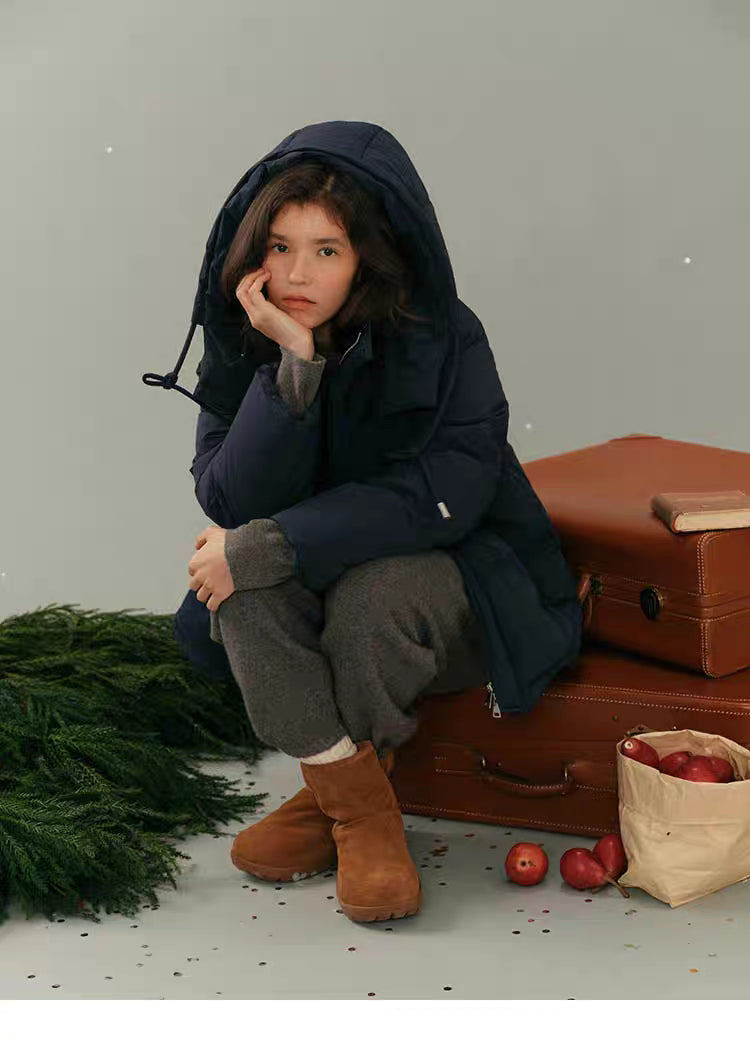 Model sitting with hood on, wearing navy blue puffer jacket and looking forward next to luggage.