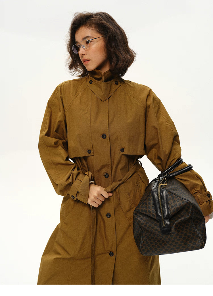 Side view of woman wearing belt-tied maple brown trench coat holding travel bag.