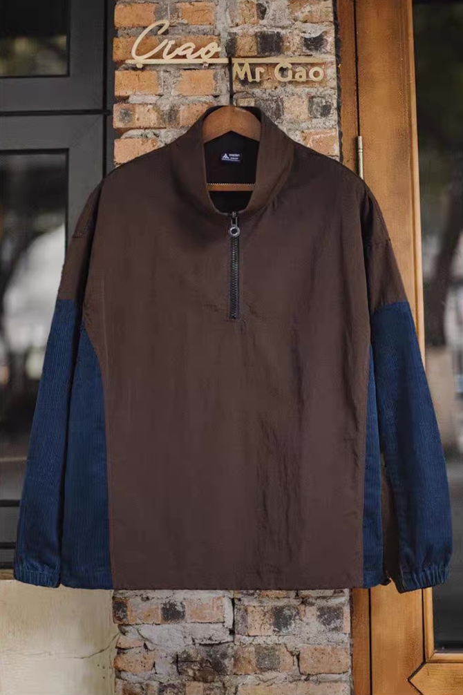 URBANCORE half-zip nylon and corduroy patchwork jacket hanging on a wooden hanger beside a brick wall, showing stand collar and casual silhouette.