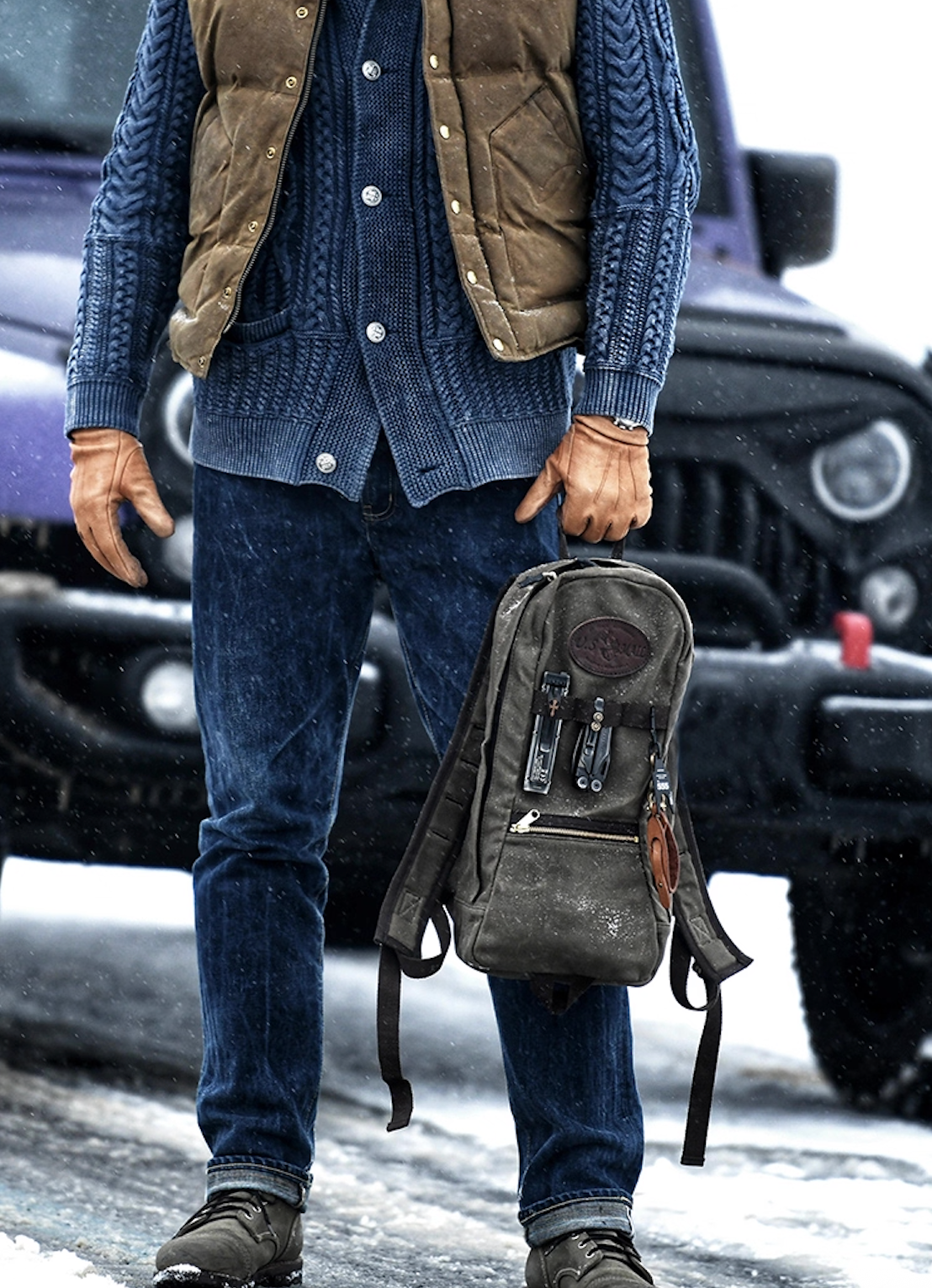 Model holding U.S. Mail waxed canvas backpack beside Jeep wearing outdoor gear.