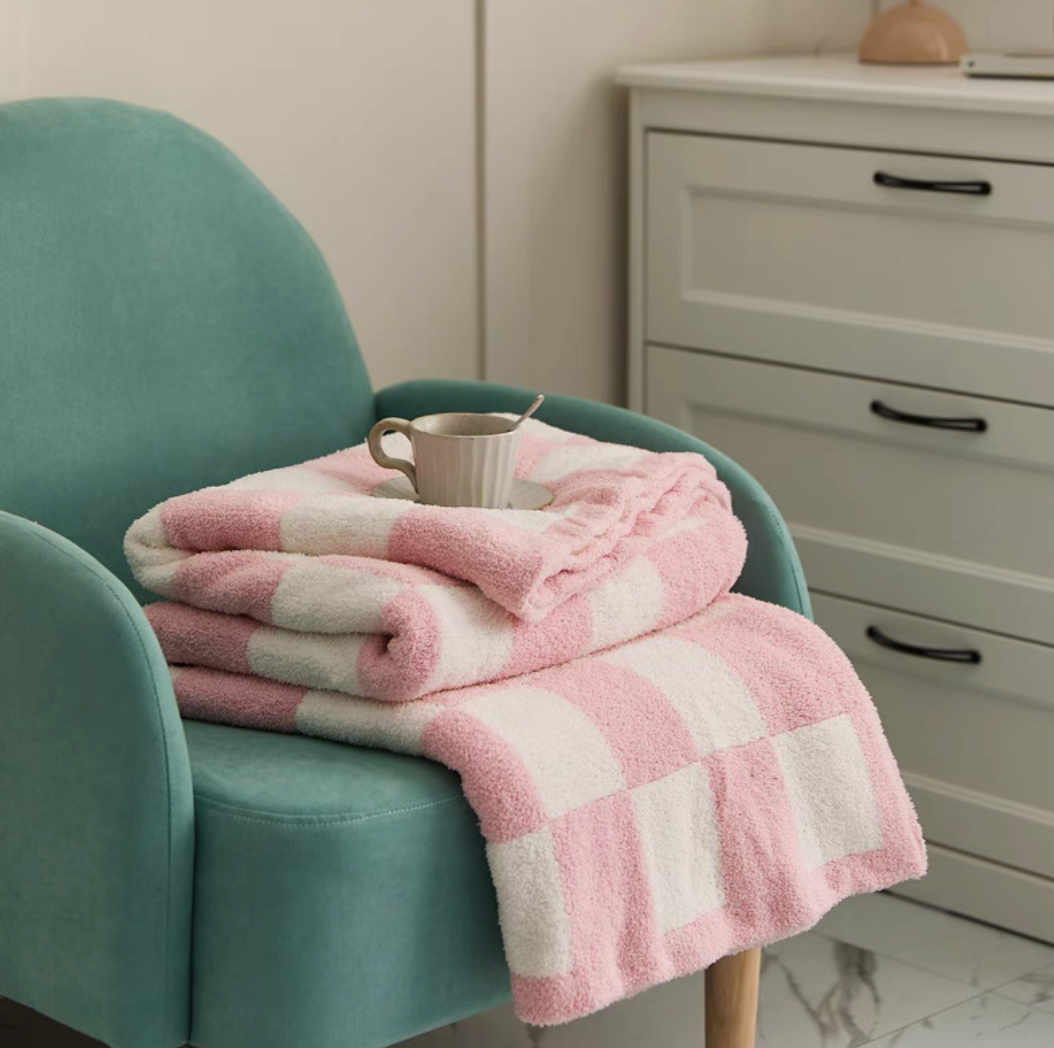Folded pink checkered blanket stacked on teal armchair with coffee cup placed on top in relaxed home setting
