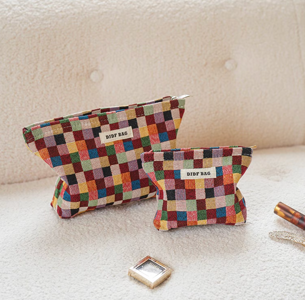 Two sizes of Vibrant Checkered Travel Makeup Bag in multi-colored checkered pattern placed on soft fabric surface with beauty accessories nearby