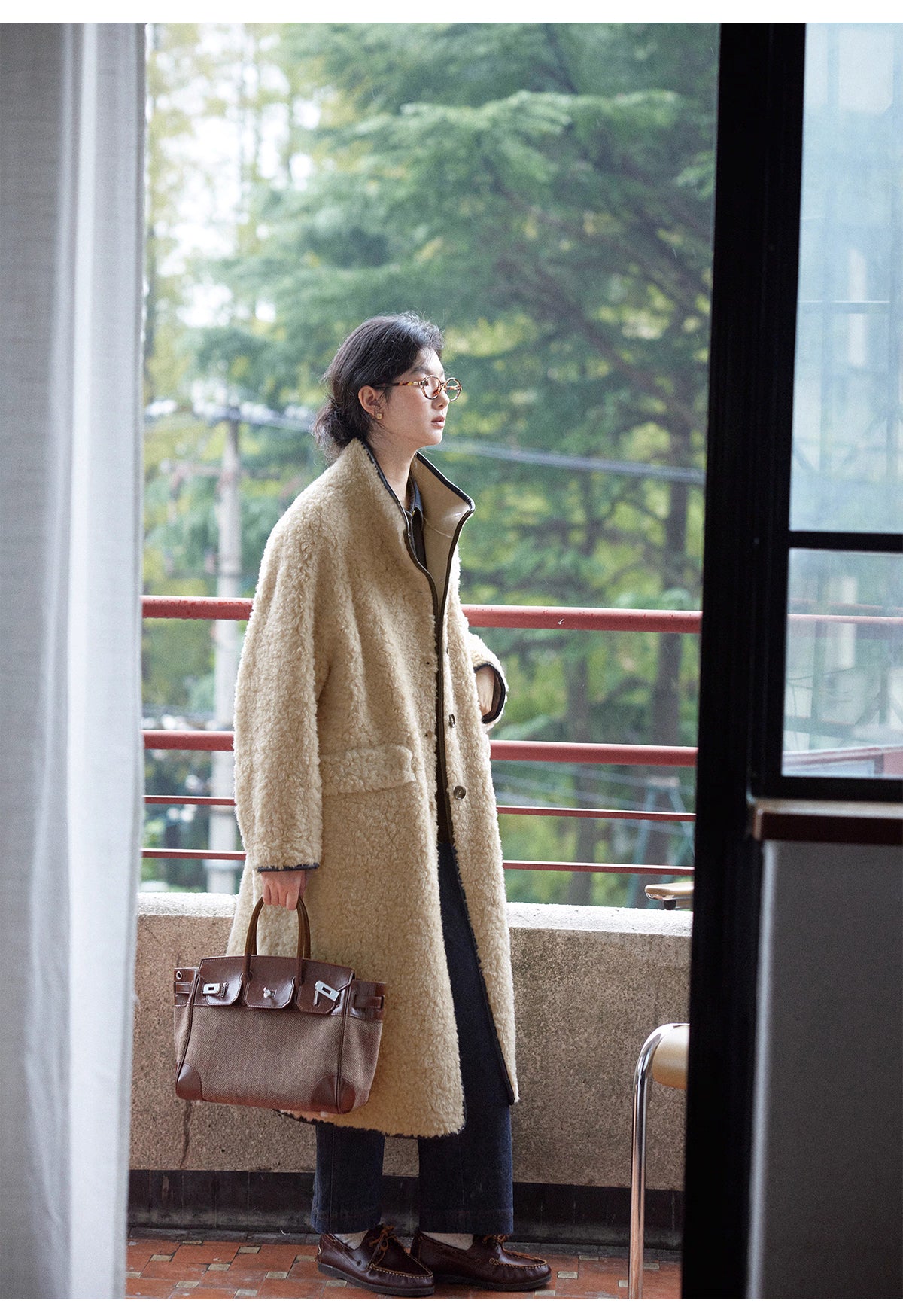 Woman wearing oversized wool shearling coat on a balcony, styled with trousers and leather tote bag.