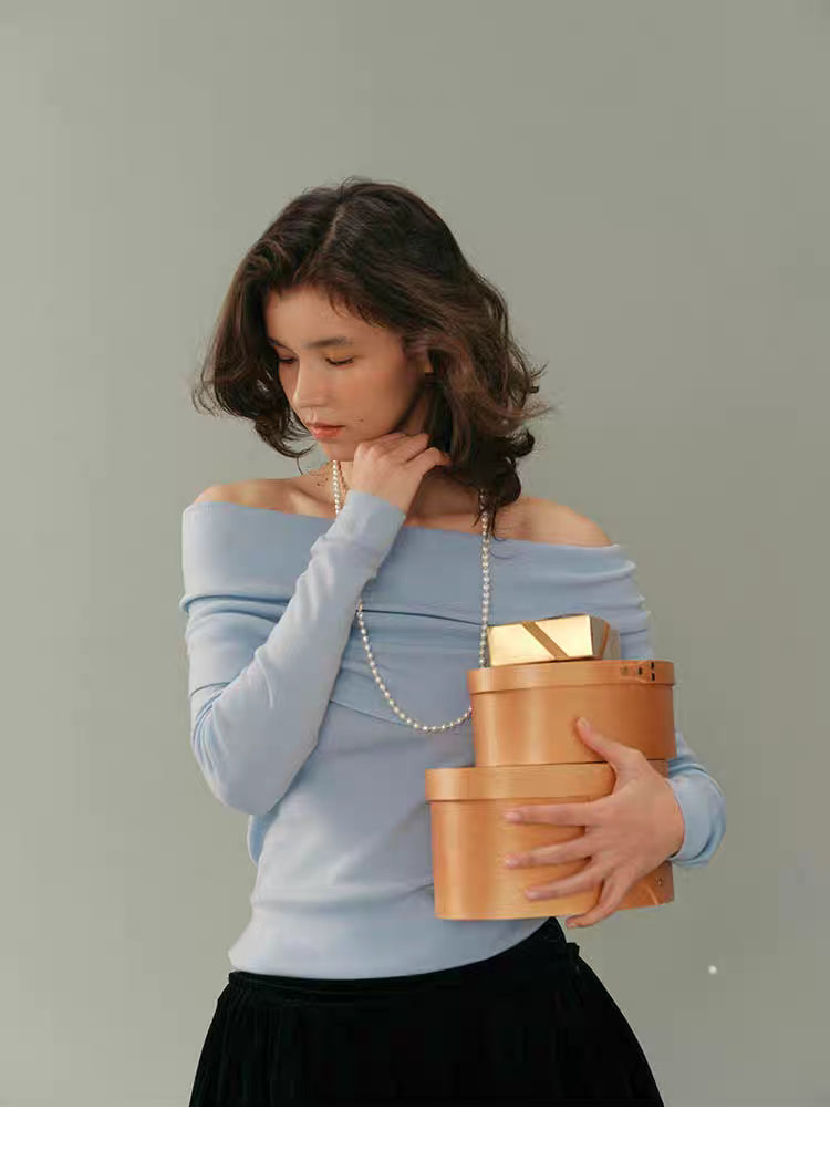 Model wearing soft sky-blue off-shoulder wool top holding stacked wooden gift boxes