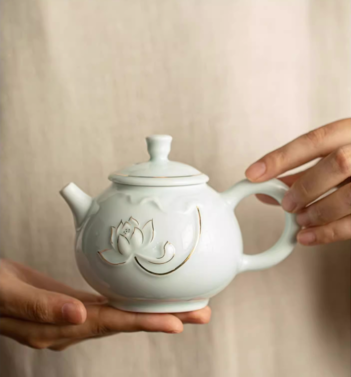 Yingqing Lotus Relief Ceramic Teapot in white color with embossed lotus design and gold detailing, front view held in hands showing rounded body and lid