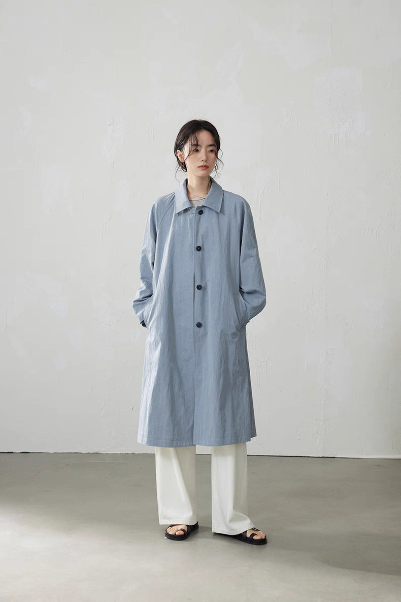 Model wearing light blue-gray A-line raglan trench coat with white pants, minimalist studio setting.