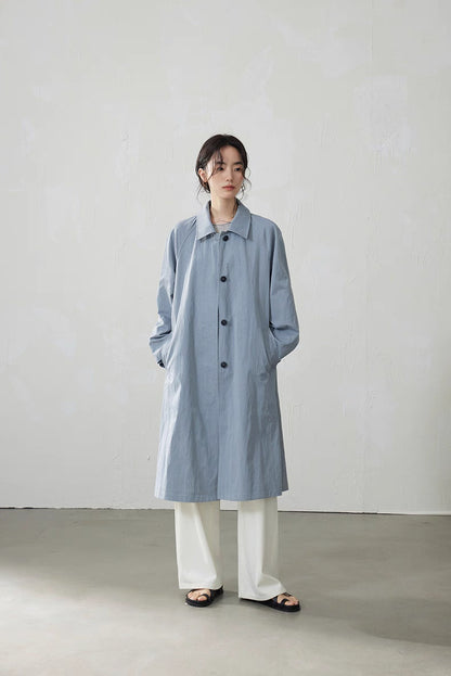 Model wearing light blue-gray A-line raglan trench coat with white pants, minimalist studio setting.