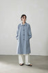 Model wearing light blue-gray A-line raglan trench coat with white pants, minimalist studio setting.