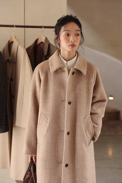 Woman wearing oat beige Suri alpaca long coat standing in studio with hanging coats behind her.