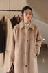 Woman wearing oat beige Suri alpaca long coat standing in studio with hanging coats behind her.