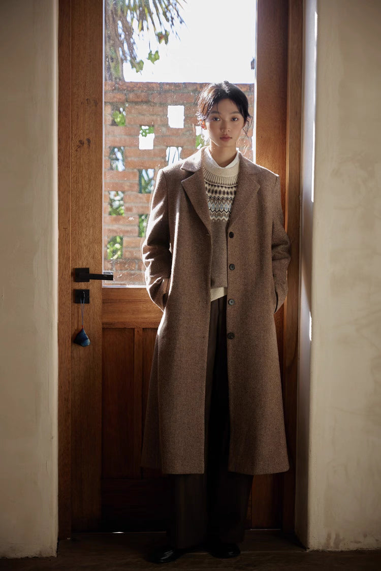 Woman wearing a brown herringbone wool long coat, standing by a wooden door in soft natural light.