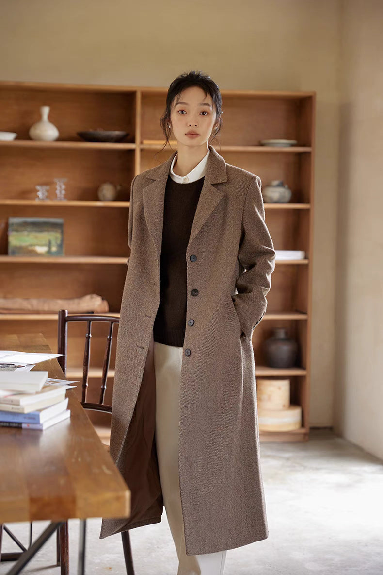 Elegant woman in a brown wool coat layered over a white shirt and dark sweater, standing in a cozy study room.