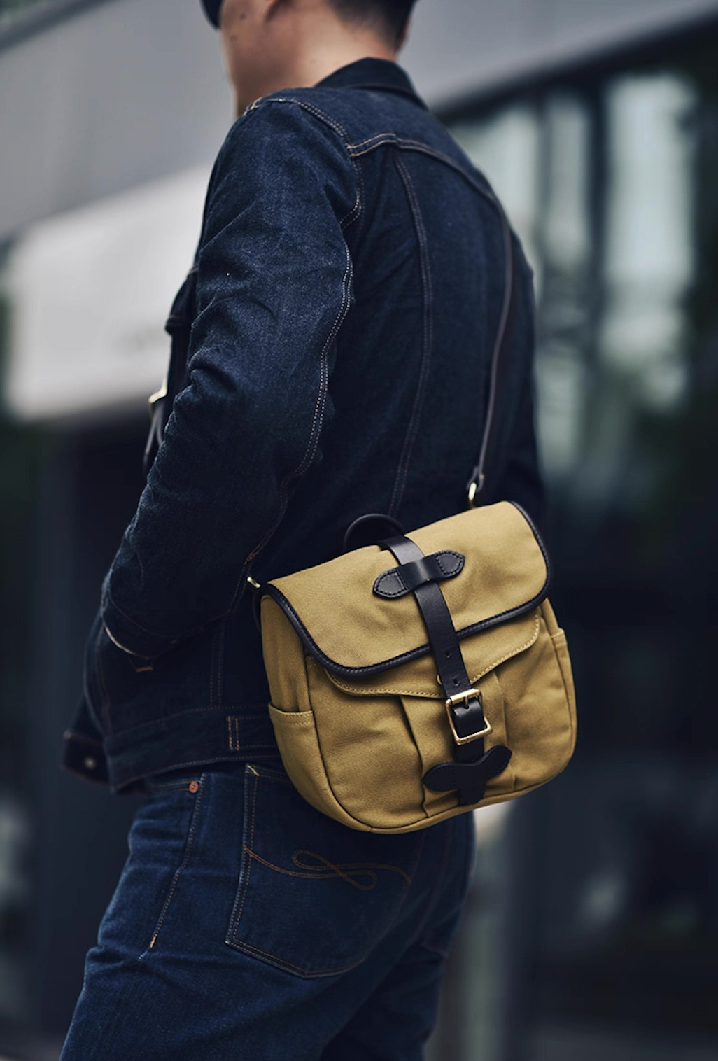 Man wearing Nomad Field Tan crossbody satchel over denim jacket outdoors.