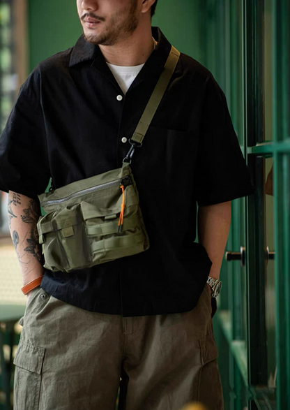 Man wearing army green crossbody bag with black shirt and khaki pants in urban setting
