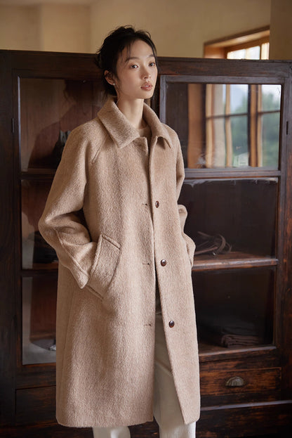 Side view of woman wearing oat beige alpaca long coat standing near wooden shelves.