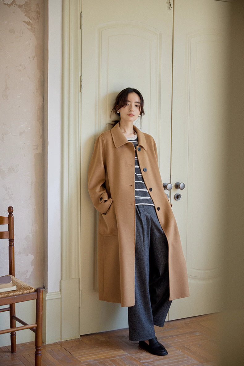 Woman leaning against wall in camel coat over striped pullover and wool trousers.