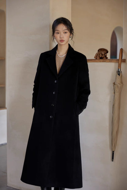 Woman in a long black wool coat with a pearl necklace, standing indoors in a minimalist beige room.