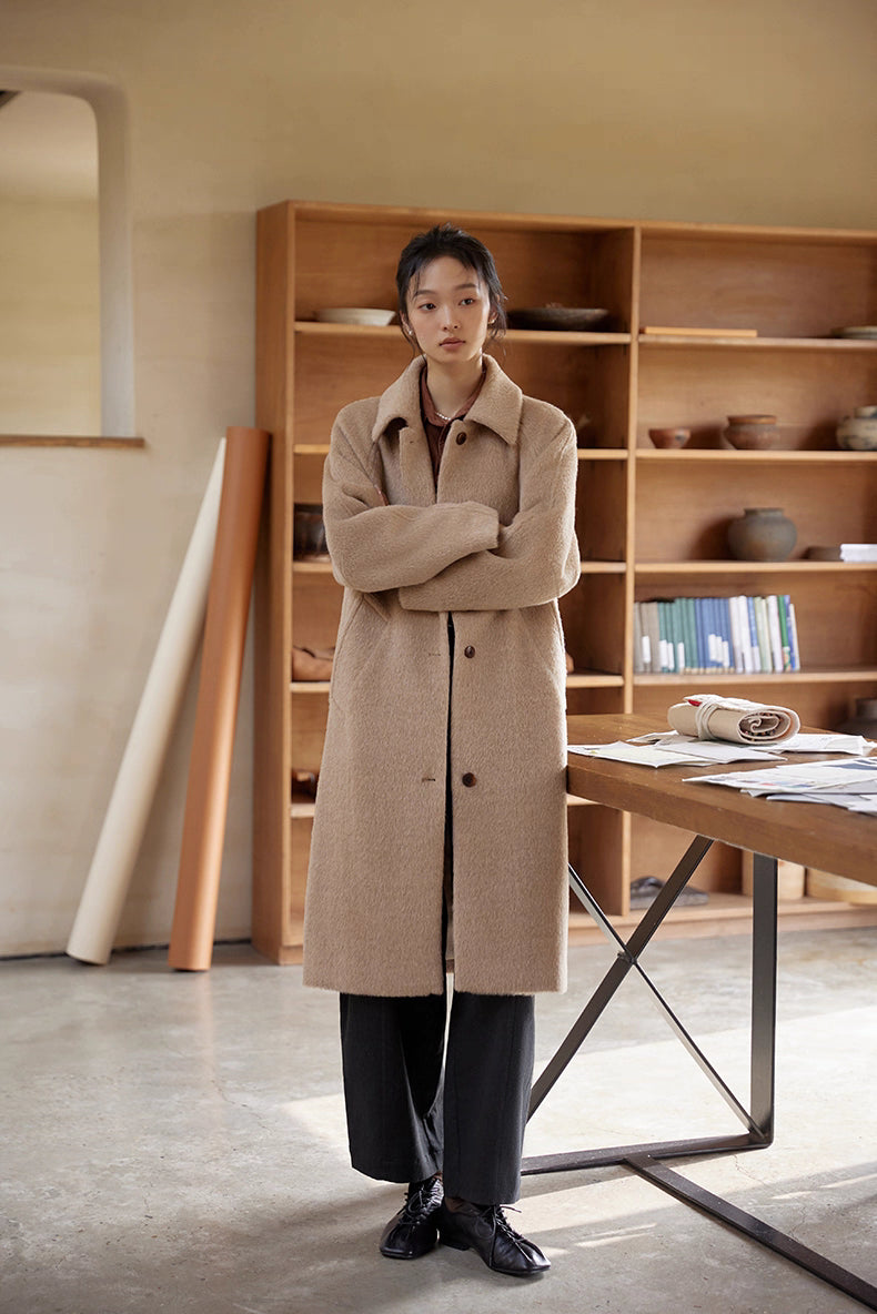 Woman crossing her arms while wearing a long oat beige alpaca wool coat in a minimal room.