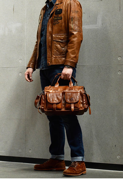 Man in denim and boots holding camel brown leather work bag in front of gray wall