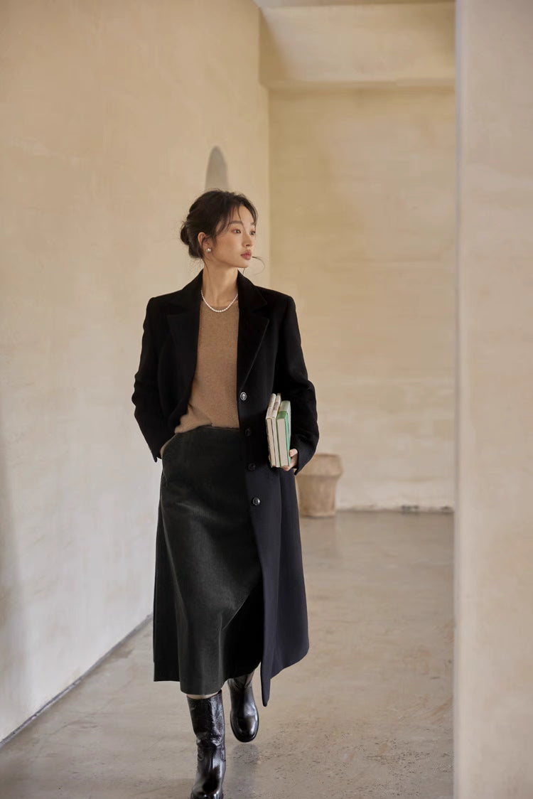 Model wearing a tailored black coat paired with a midi skirt and boots, holding books while walking indoors.