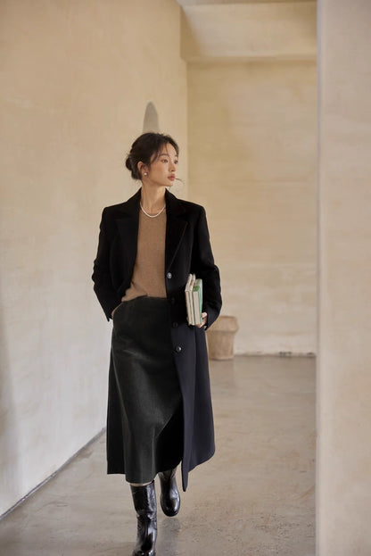 Model wearing a tailored black coat paired with a midi skirt and boots, holding books while walking indoors.