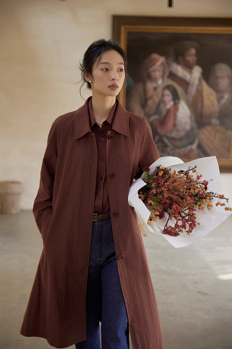 Model wearing terracotta brown A-line raglan trench coat with neutral drape backdrop.