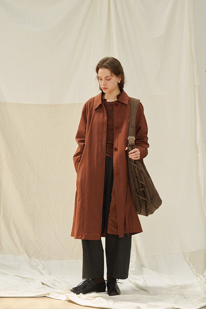 Front view of terracotta brown trench coat styled with layered brown top and gray trousers.