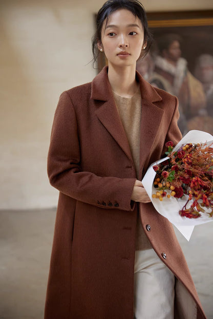 Woman in a warm brown wool long coat holding a bouquet of autumn flowers in a soft indoor setting.