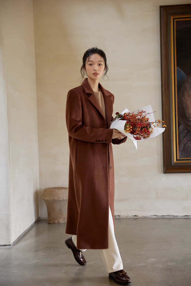 Woman wearing a brown wool coat paired with beige sweater and white pants, holding a bouquet while walking indoors.