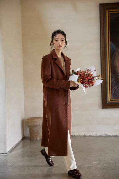 Woman wearing a brown wool coat paired with beige sweater and white pants, holding a bouquet while walking indoors.