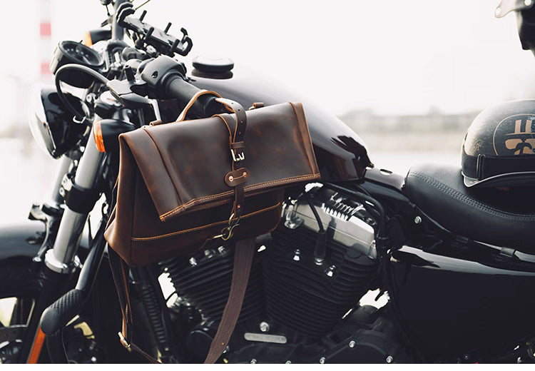 Handcrafted brown leather messenger bag displayed on a motorcycle beside a helmet, highlighting its durable design and adventurous style.