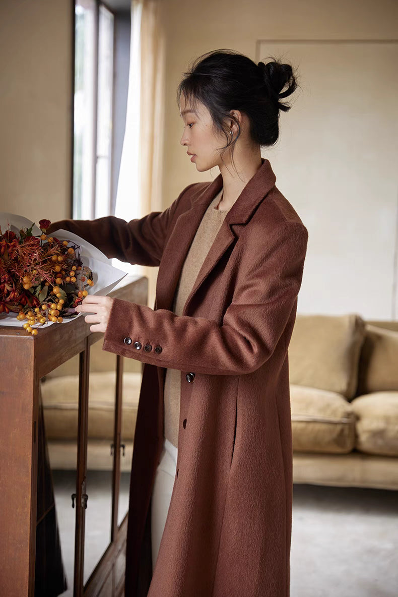 Side profile of a woman arranging flowers in a vase while wearing a brown wool long coat in a cozy living space.