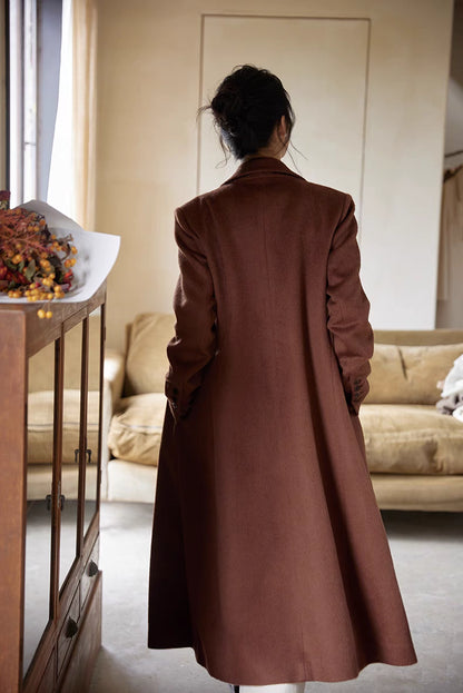 Back view of a woman in a brown wool long coat standing near a wooden cabinet with flowers in a calm interior.