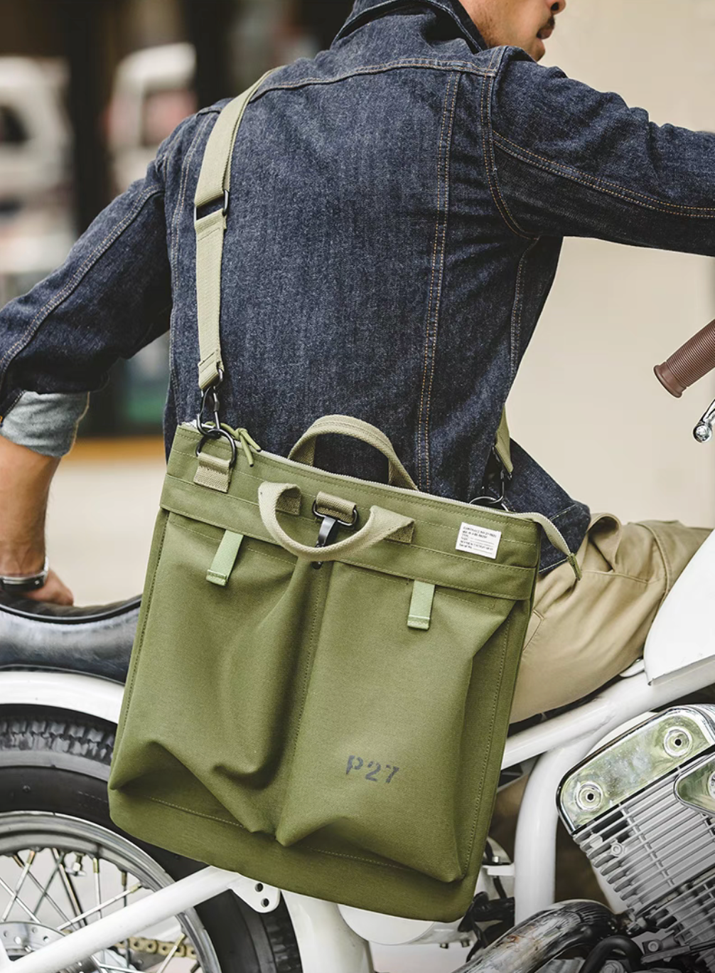 Person wearing army green canvas tote bag crossbody while sitting on a motorcycle, demonstrating adjustable strap use.