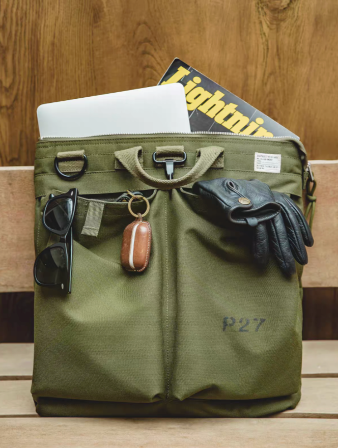Army green canvas tote bag with front pockets holding sunglasses, key holder, and gloves, with a laptop and magazine visible inside on a wooden bench.