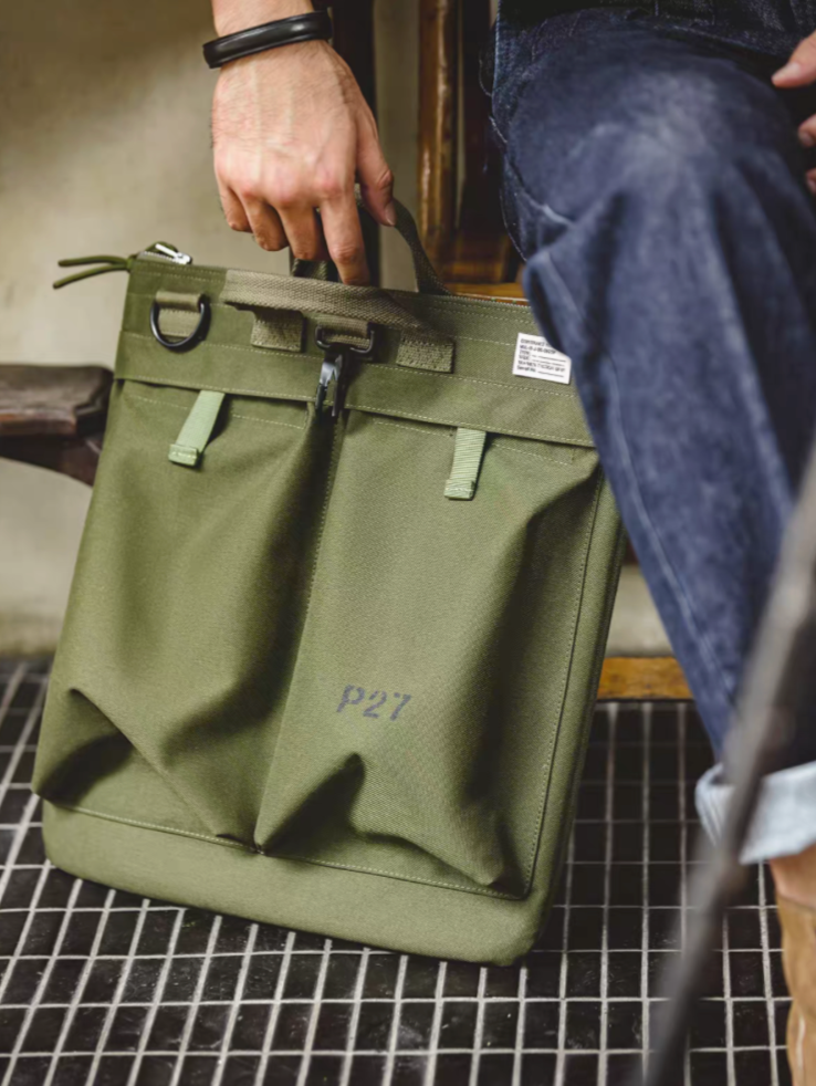 Person holding army green canvas tote bag by the top handles, showing front pockets and structured design in a casual indoor setting.