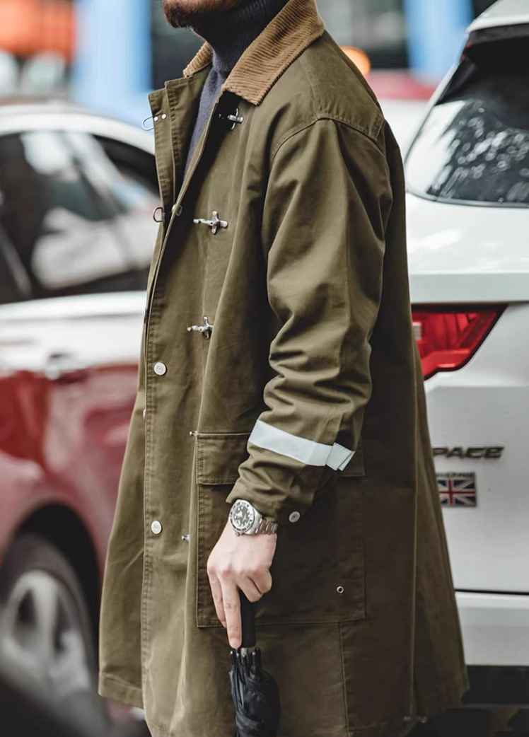 Man wearing olive green workwear coat with toggle closure and reflective sleeve detail, urban street style.