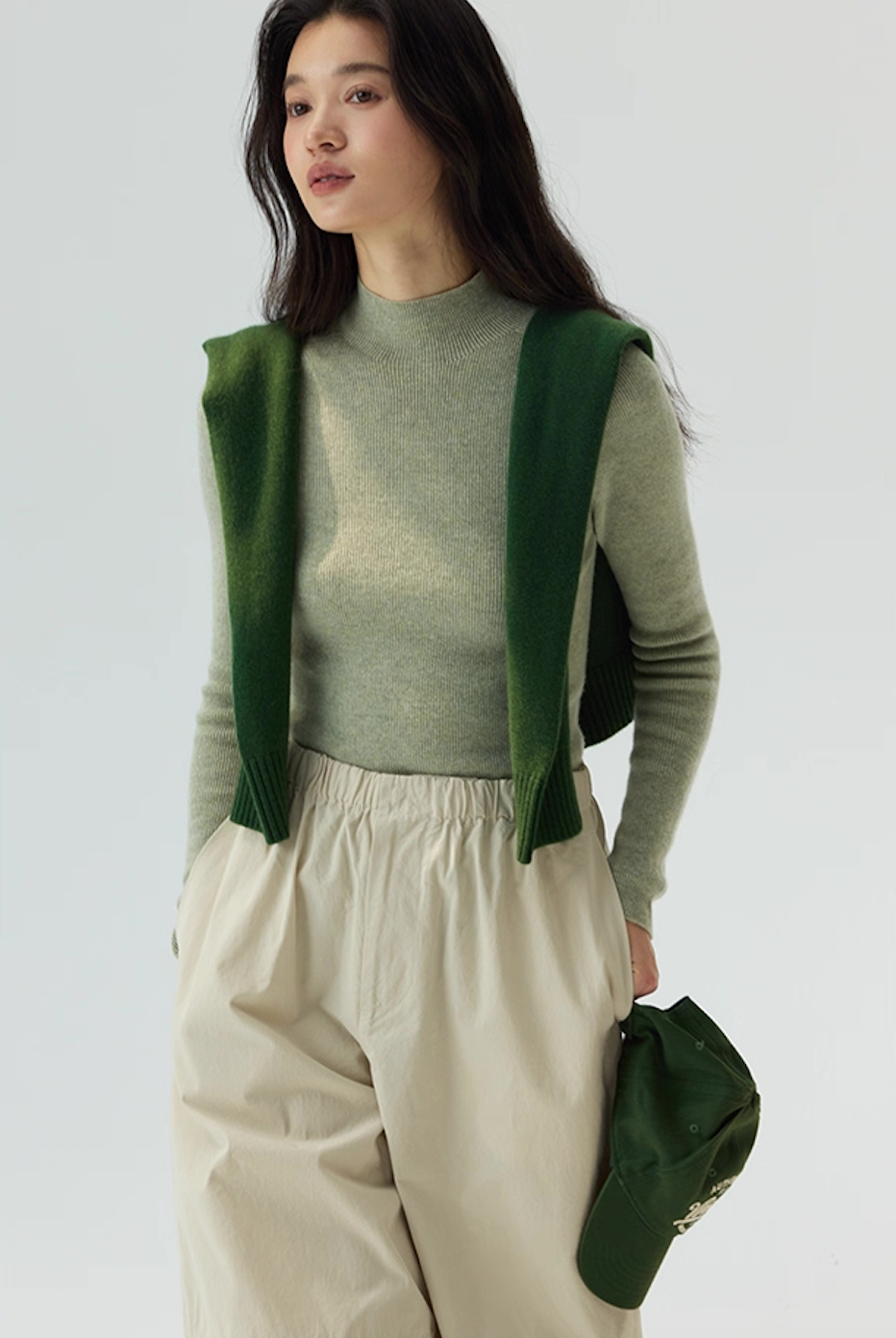 Woman in sage green cashmere sweater with forest green knit over shoulders, paired with beige pants