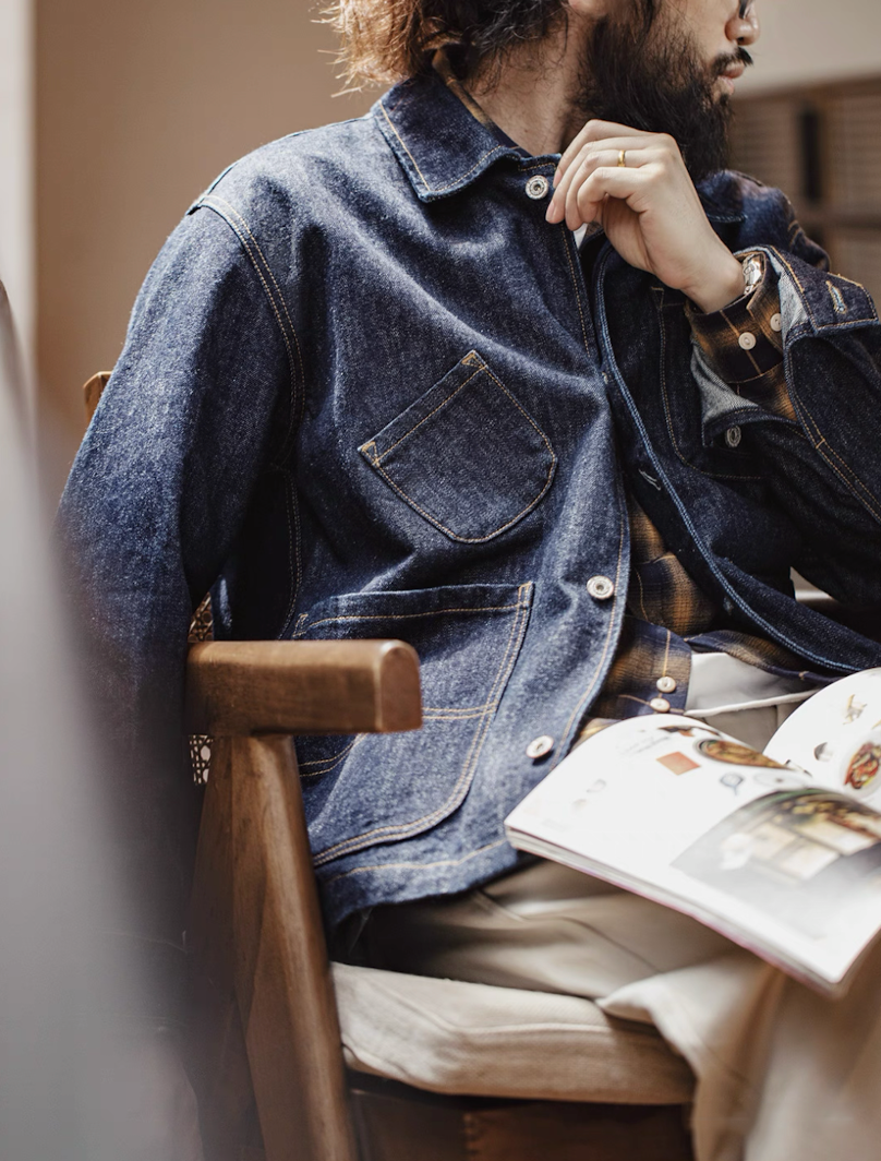 Man sitting in chair wearing 14 oz denim utility jacket reading a magazine