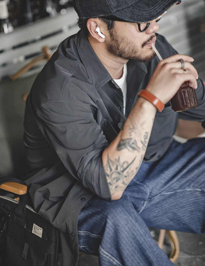 Model sitting in café, wearing men’s dark charcoal overshirt jacket with rolled-up sleeves.