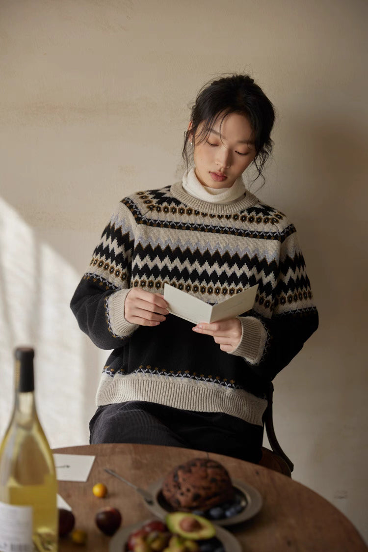 Model in black-cream Fair Isle sweater reading at wooden table with fruit and wine