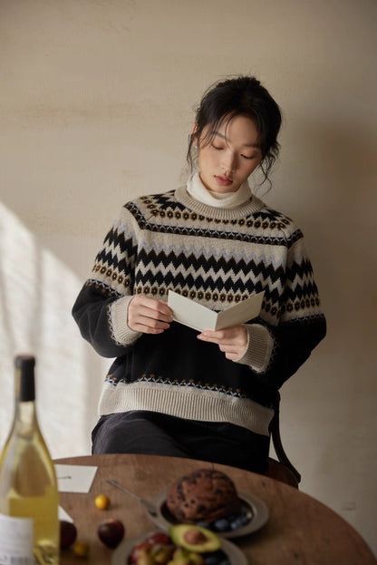 Model in black-cream Fair Isle sweater reading at wooden table with fruit and wine