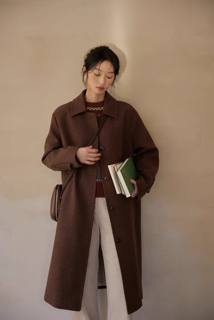 Model leaning by wall in brown coat over gray-red Fair Isle sweater holding books