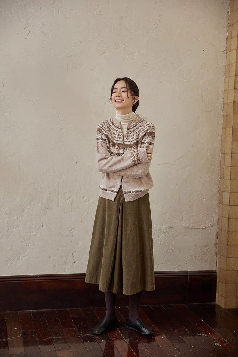 Full-body view of model in beige-brown Fair Isle cardigan and olive skirt standing by wall