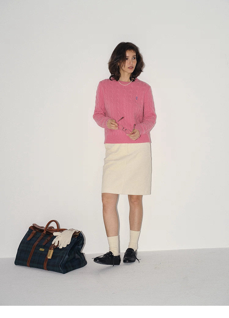 Full-body shot of a woman in a rose pink pure wool cable-knit sweater paired with a cream skirt, standing beside a plaid weekender bag in studio.