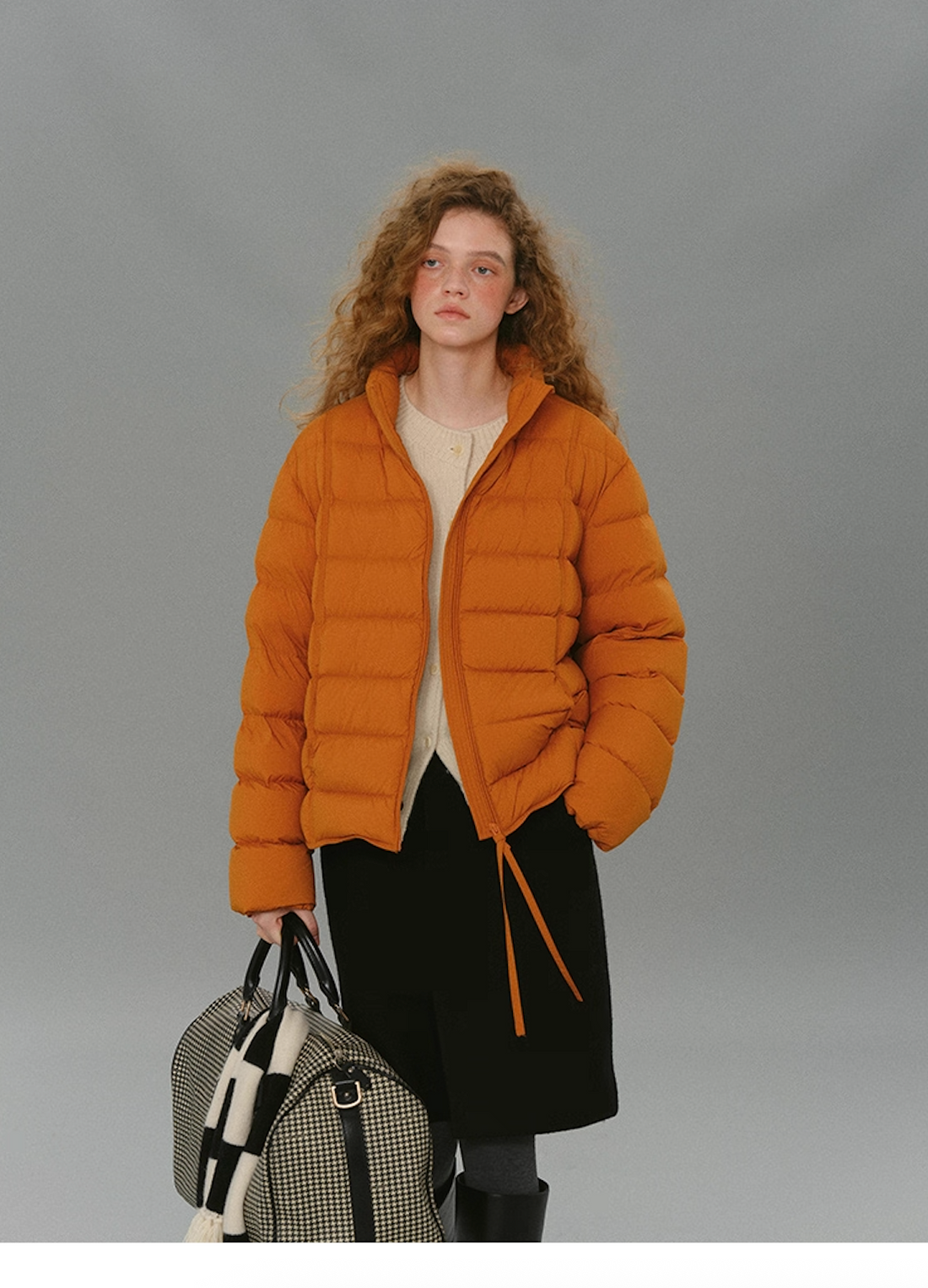 Model wearing a warm orange matte down puffer holding a checkered duffle bag.