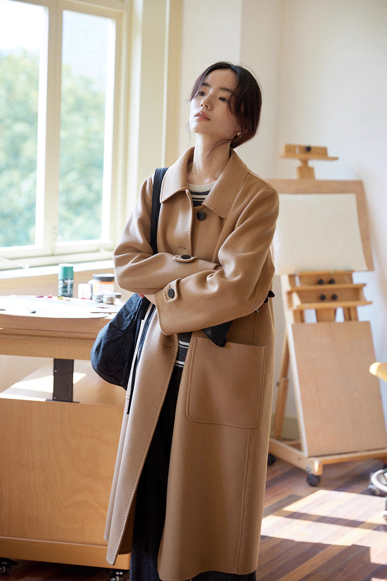Almond-camel cashmere wool long coat styled with striped sweater, model standing with arms crossed in an art studio